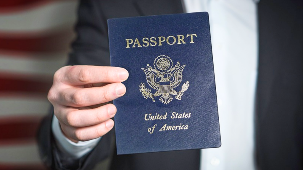 A person wearing a dark suit holds up a United States passport with an American flag blurred in the background.