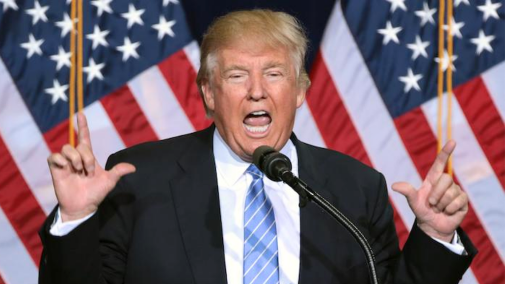 Donald Trump speaking passionately into a microphone at a podium with two American flags behind him.