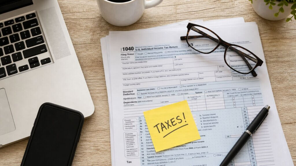 Desk with a "1040 U.S. Individual Income Tax Return" form, glasses, pen, laptop, and a sticky note reading "TAXES!", symbolizing the financial focus behind the silicon six tax issue.