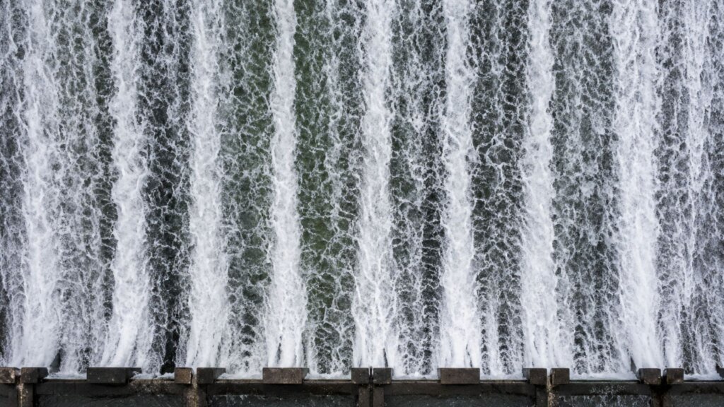 White water rushing over a dam in evenly spaced vertical streams viewed from above. Dark concrete barriers line the bottom edge beneath the flowing water.