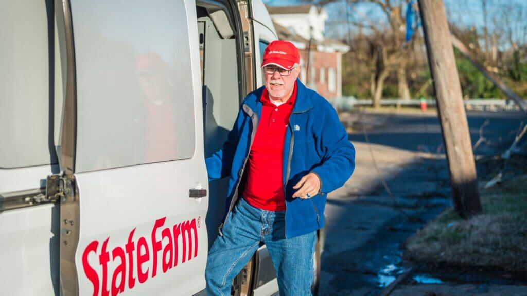 State Farm worker in red shirt and hat stepping out of a branded van in a residential area, representing insurance service response in the community.
