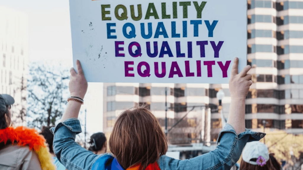 A low-angle shot from behind a protestor holding up a white sign that reads "EQUALITY" repeated four times in green, blue, purple, and magenta.