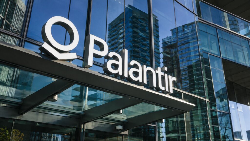 Large Palantir company logo mounted on the glass exterior of a modern office building with reflective windows and surrounding city skyscrapers.