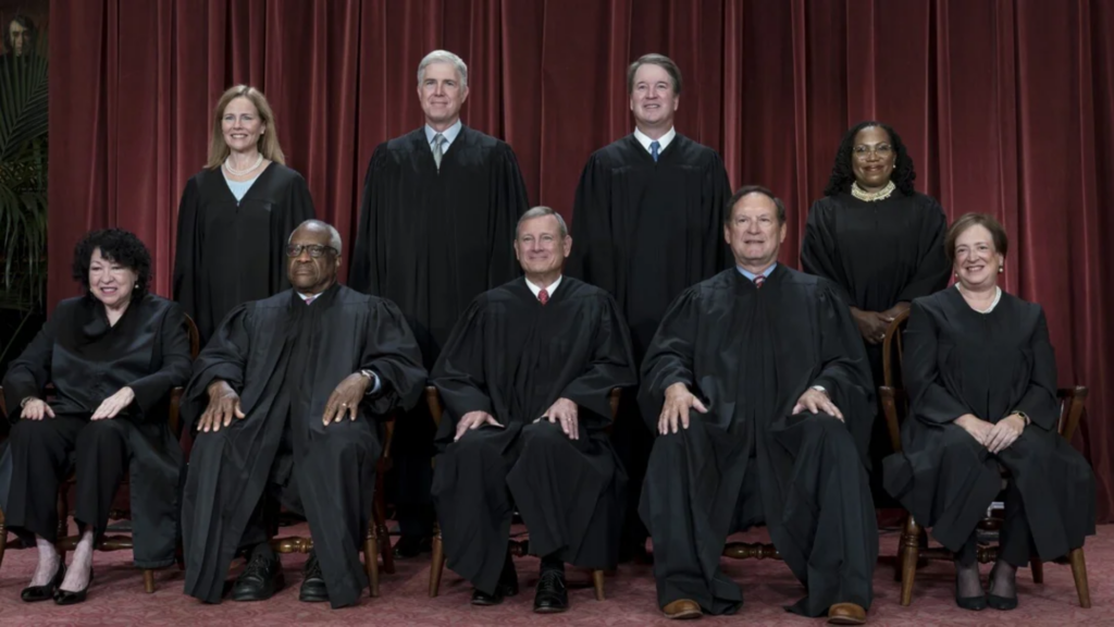 A formal group portrait of the nine U.S. Supreme Court Justices wearing their black judicial robes, arranged in two rows against a backdrop of deep red curtains.