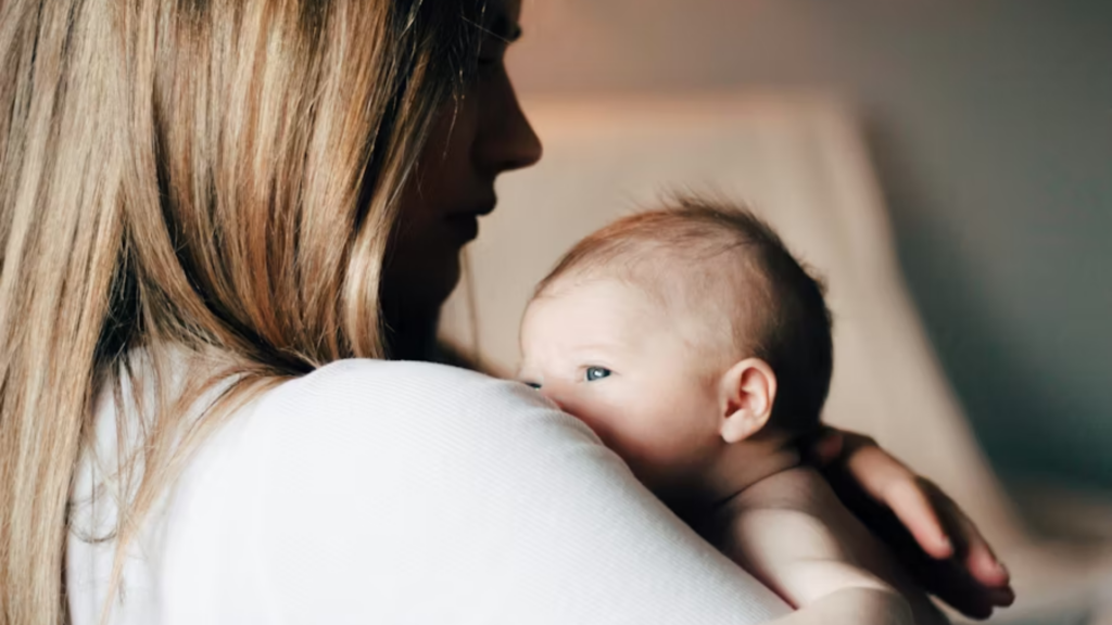 A tender, silhouetted profile shot of a woman with long blonde hair holding a newborn baby against her shoulder; the soft lighting emphasizes the emotional bond and the "skin-to-skin" contact during recovery.