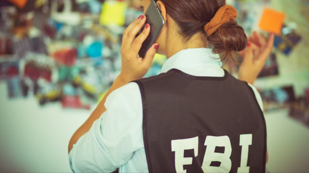 An over-the-shoulder shot of a woman with her hair in a bun, talking on a smartphone; she is wearing a black tactical vest with bold white "FBI" lettering, standing in front of a blurred investigation board covered in notes and photos.