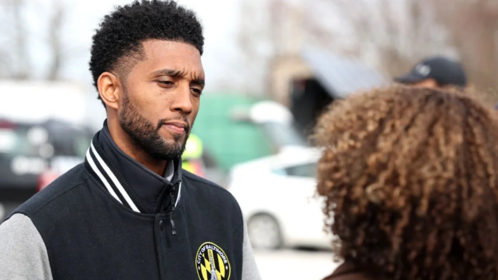 A medium shot of Baltimore Mayor Brandon Scott wearing a black-and-grey varsity jacket adorned with the official circular "City of Baltimore" seal.