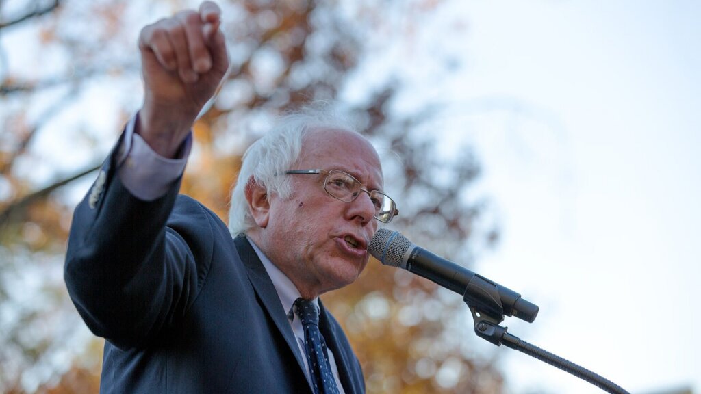 Sen. Bernie Sanders, in a suit speaks into a microphone outdoors while raising his fist during a public speech or political rally.