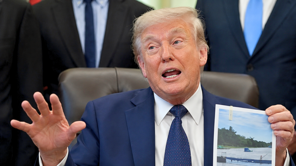 A close-up portrait of Donald Trump speaking animatedly during an Oval Office briefing; he wears a blue suit and dotted blue tie, gesturing with an open right hand while his left hand holds up an aerial photograph displaying the empty, gray stone channel of the reflecting pool undergoing initial draining and staging.