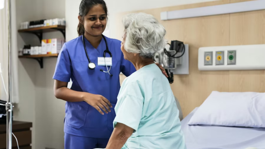 A horizontal medical shot showing a young female nurse or doctor in blue scrubs and a stethoscope smiling warmly at an elderly female hospital patient in a light green gown, resting a hand gently on her shoulder in a modern hospital room.