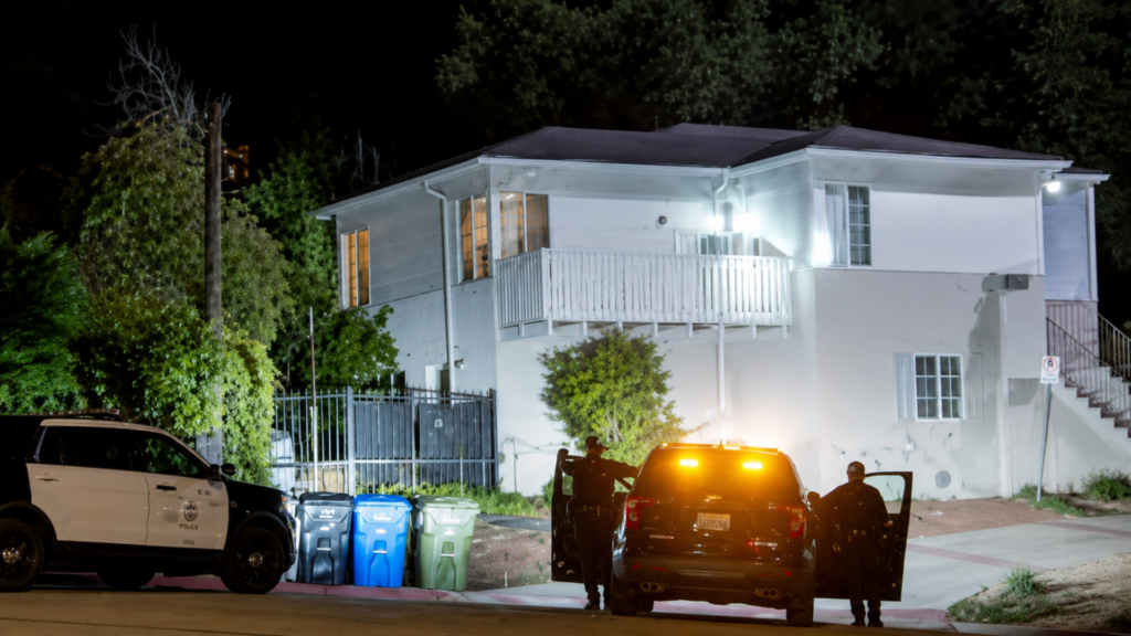 A nighttime shot of a residential street outside a white two-story house; a black-and-white police cruiser is parked on the left, while two police officers stand near the open doors of an SUV with its yellow emergency flashers activated.