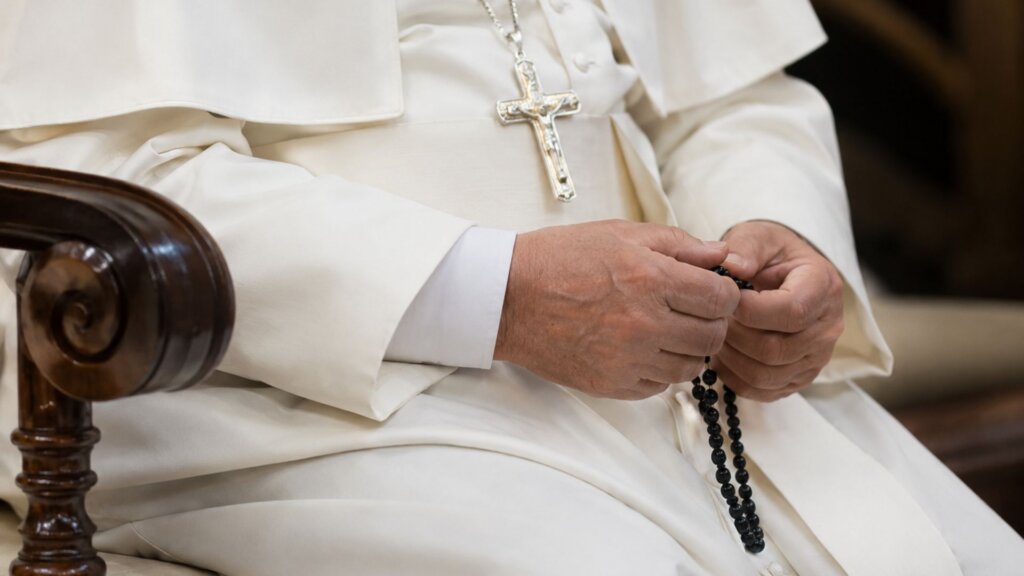 Pope sits in white papal robes holding a black rosary in both hands, with a silver cross hanging over his chest.
