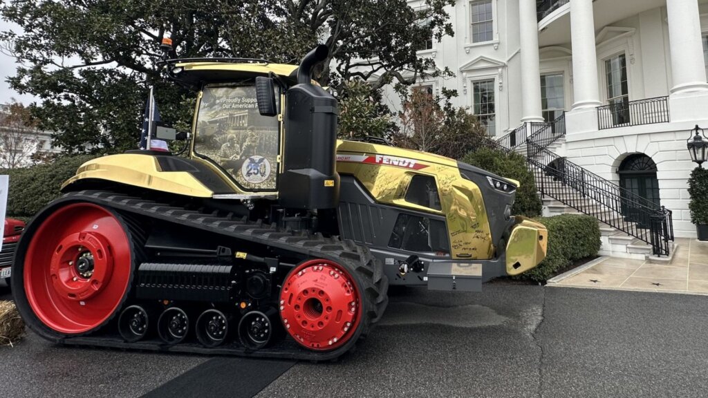 Large gold and black tractor with red wheels parked outside a white government building. The tractor is covered with signatures and displays the text “Proudly Supporting Our American Farmers,” highlighting the connection between farming interests and national policy.