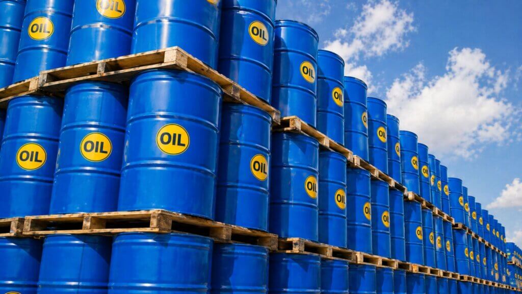Rows of stacked blue oil barrels on wooden pallets outdoors beneath a partly cloudy sky. Each barrel has a yellow circle labeled “OIL” printed on the side.