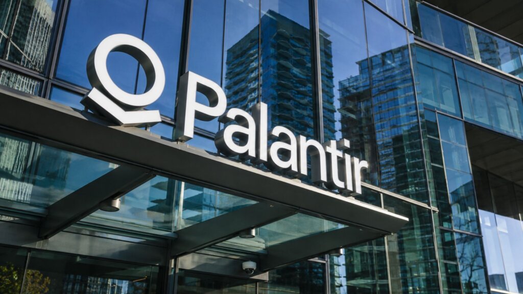 “Palantir” sign mounted above the entrance of a modern glass office building with reflections of nearby skyscrapers in the windows.