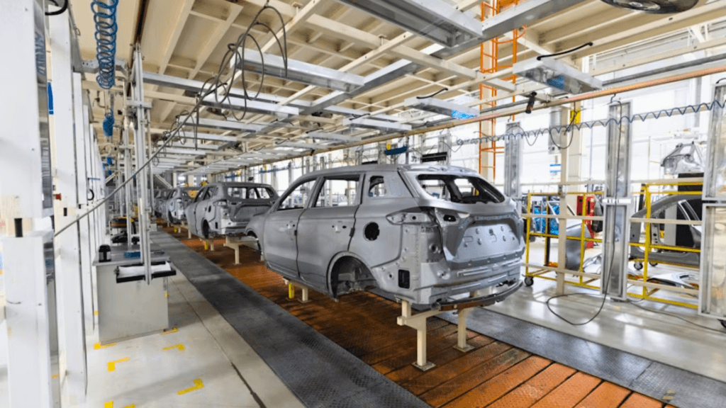 A wide-angle shot of a modern automotive assembly line; several unpainted grey SUV or car bodies are mounted on yellow platforms along a moving track, surrounded by overhead robotic arms and industrial lighting.