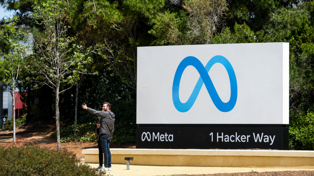 A wide daytime shot of the iconic 1 Hacker Way sign at Meta's headquarters, featuring the blue infinity-loop logo; a couple is standing in the foreground taking a selfie in front of the sign, surrounded by green trees.