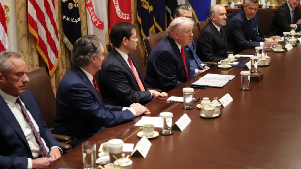 A wide high-angle shot of a White House Cabinet Room meeting; President Trump sits at the center of a long dark-wood conference table next to Florida Senator Marco Rubio (currently serving as Secretary of State), surrounded by various advisers, military leaders, and coffee cups.
