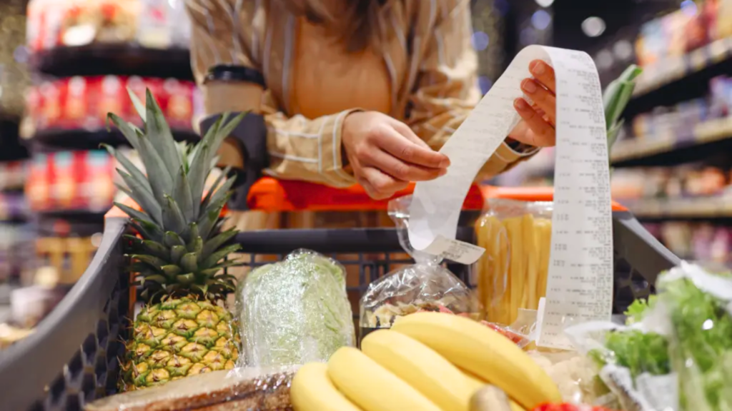 A person looking over a long receipt next to a shopping cart filled with fresh produce like a pineapple and bananas.