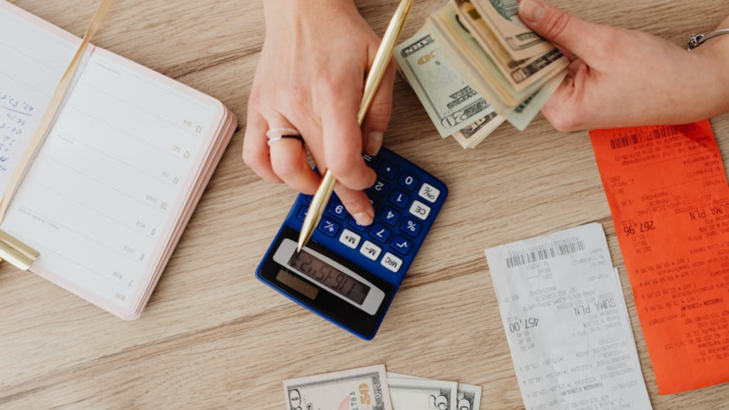 An overhead view of hands counting dollar bills and using a calculator on a wooden desk next to store receipts.