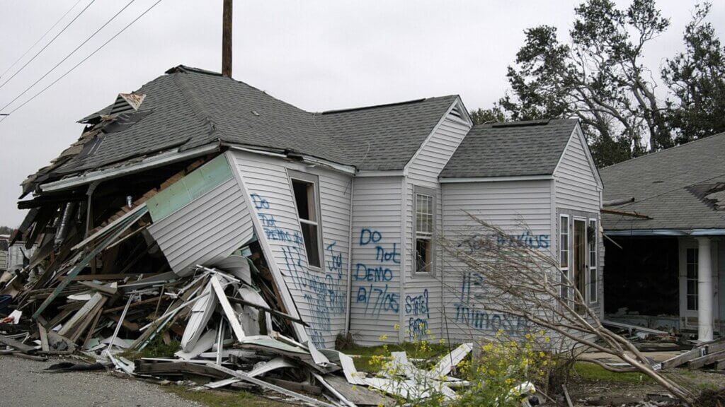 Severely damaged house with collapsed walls and debris scattered outside, with spray painted warning text including "DO NOT DEMO 9/7/05" highlighting destruction tied to insurance claims and allegations.