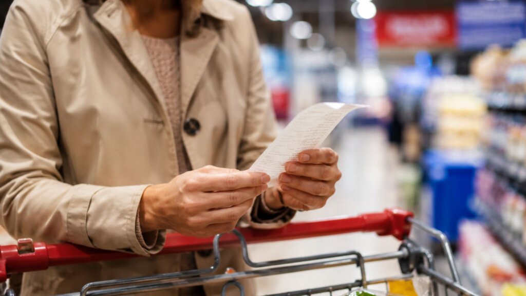 Shopper in a grocery store stands beside a shopping cart while holding and reading a long printed receipt. Brightly lit store aisles and shelves appear blurred in the background.
