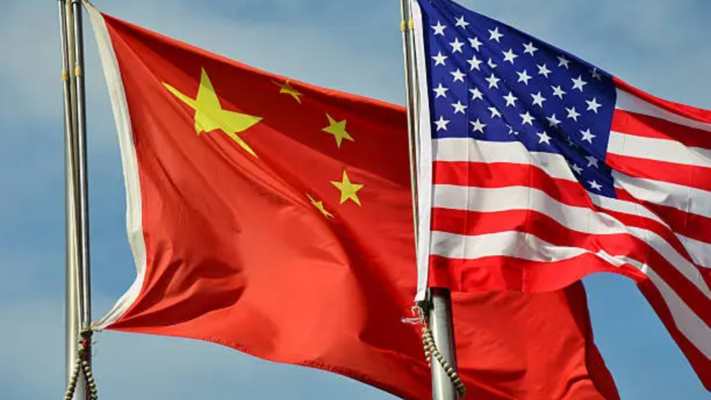 A clear daytime shot of two flags flying on metal poles against a hazy blue sky; the Chinese flag is positioned on the left, while the American flag flies on the right.