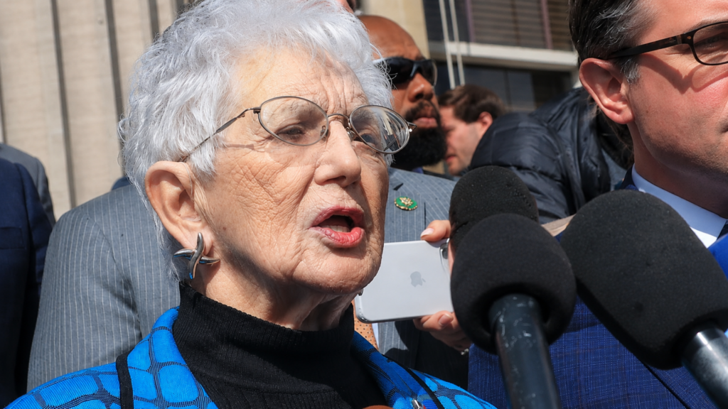A close-up, high-contrast shot of Virginia Foxx speaking passionately into a cluster of microphones during an outdoor press conference; her expression is intense and focused.