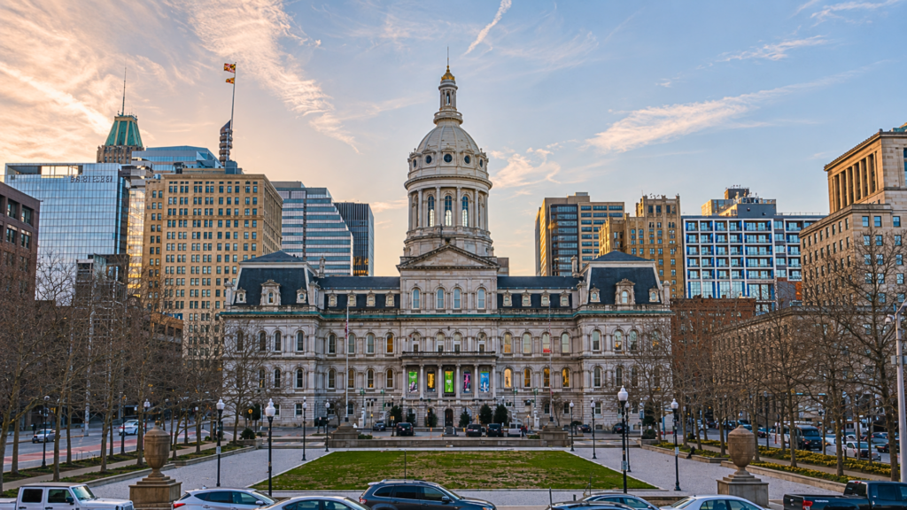 A wide daytime shot of the majestic, historic Baltimore City Hall building; the elaborate, 19th-century white building features an iconic, towering central dome rising high above a clear sky, overlooking a manicured green lawn and cars parked in front.