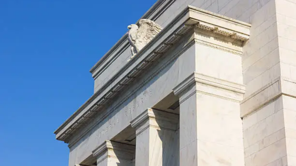 A low-angle architectural shot of the historic, white marble facade of the Federal Reserve building in Washington, D.C.; the iconic carved American eagle standard is visible along the top architrave against a clear blue sky.
