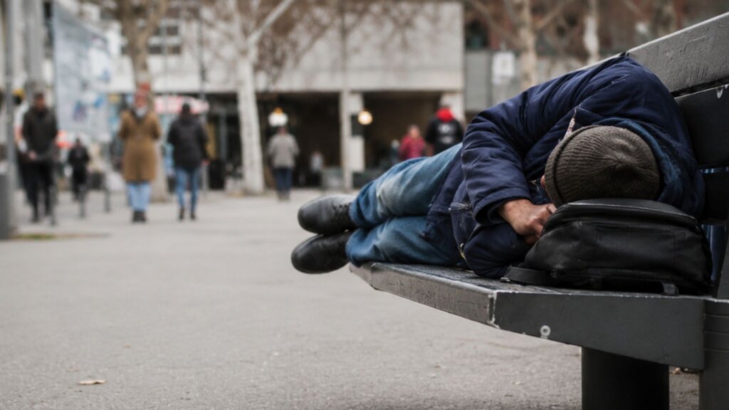 Person sleeping on a public bench in an urban area while pedestrians walk past in the background. The photo illustrates the growing homelessness crisis affecting vulnerable students and families seeking safe places to stay overnight.