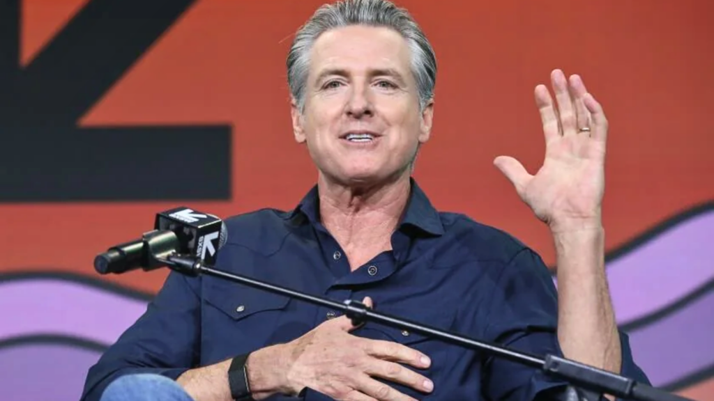 A vibrant medium shot of California Governor Gavin Newsom speaking animatedly at a panel event; he wears a navy blue button-down shirt and gestures with an open left hand while resting his right hand near his chest against an abstract orange-and-purple backdrop.