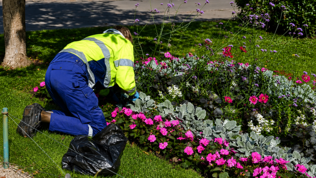 A high-angle exterior shot of a homeowner in a bright yellow reflective safety jacket and blue pants kneeling on a green lawn, looking for hidden devices.