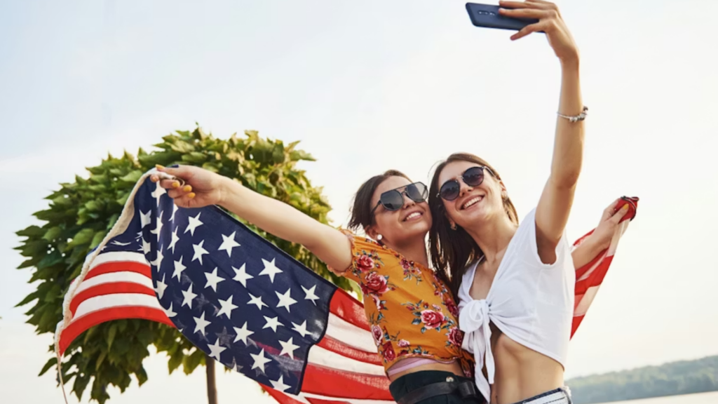 Two smiling young women in sunglasses holding up an American flag while posing to take a selfie with a smartphone outdoors.