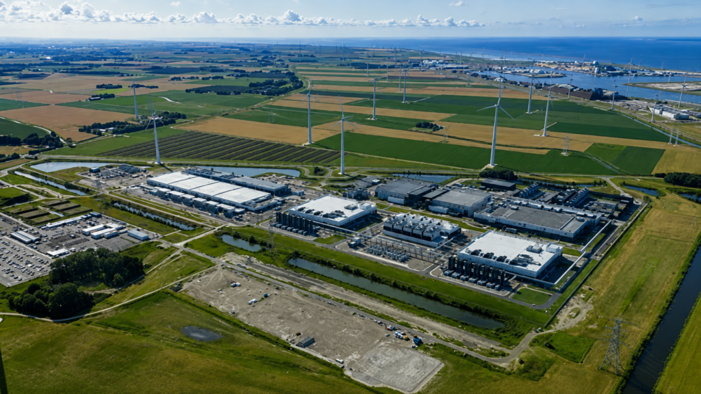 A wide aerial panoramic shot of a coastal hyperscale data center campus surrounded by farmland, green fields, and white wind turbines stretching toward the ocean.
