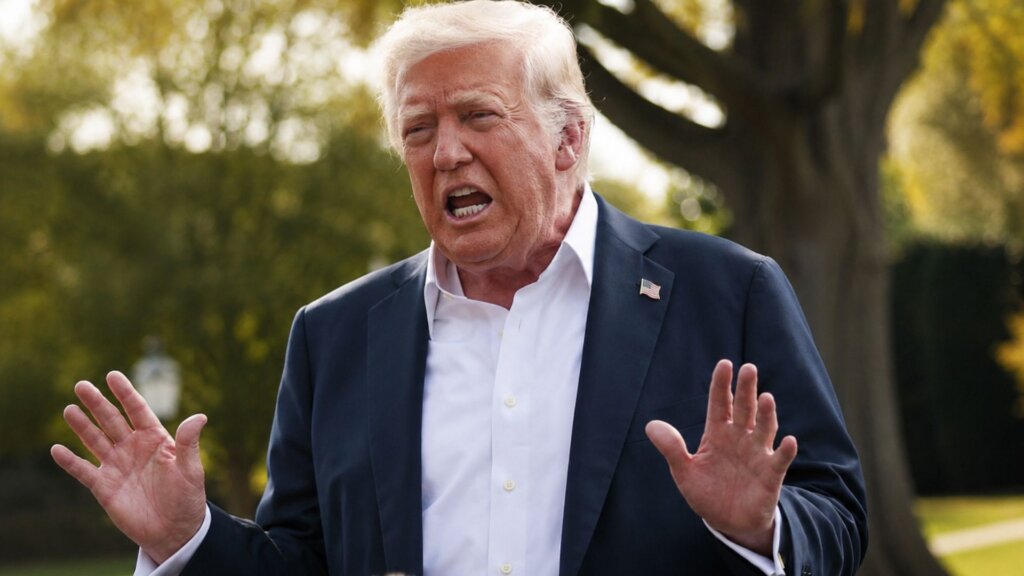 President Trump, in a dark suit and white shirt, speaks outdoors with both hands raised while standing near large trees.