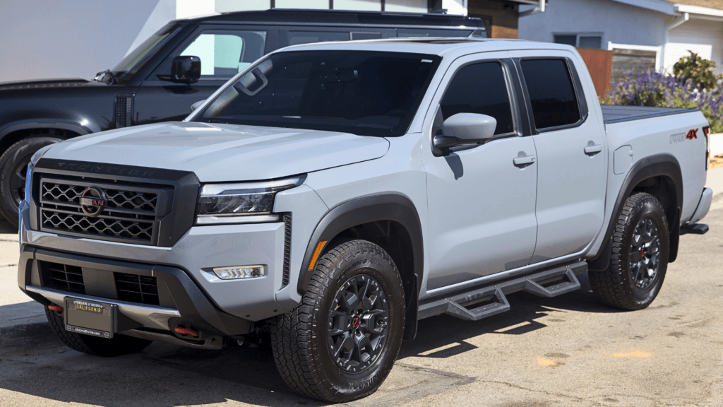 A professional-style shot of a light grey 2026 Nissan Frontier PRO-4X pickup truck parked on a residential street; the truck features black off-road wheels and a "PRO-4X" decal on the rear bed, showcasing the model's rugged design.