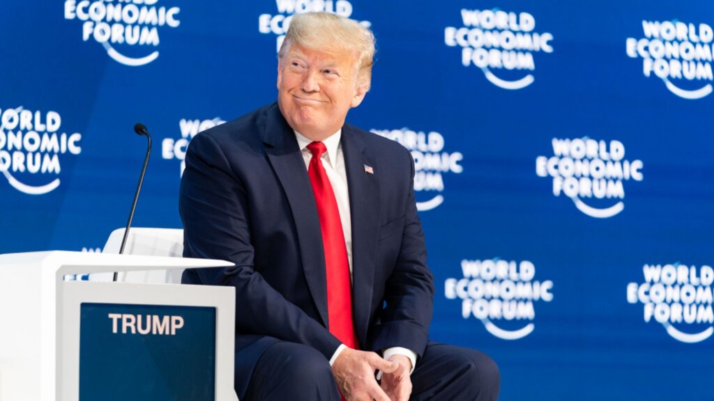 Donald Trump seated on a stage in front of a blue “World Economic Forum” backdrop wearing a dark suit with a bright red tie and smiling while looking to the side.
