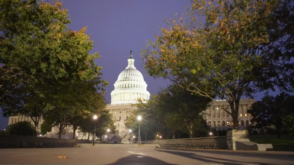 A scenic wide shot of the U.S. Capitol Building dome glowing white at twilight; the neoclassical architecture is framed by lush, leafy trees with autumn colors, overlooking a quiet, low-angle pathway.
