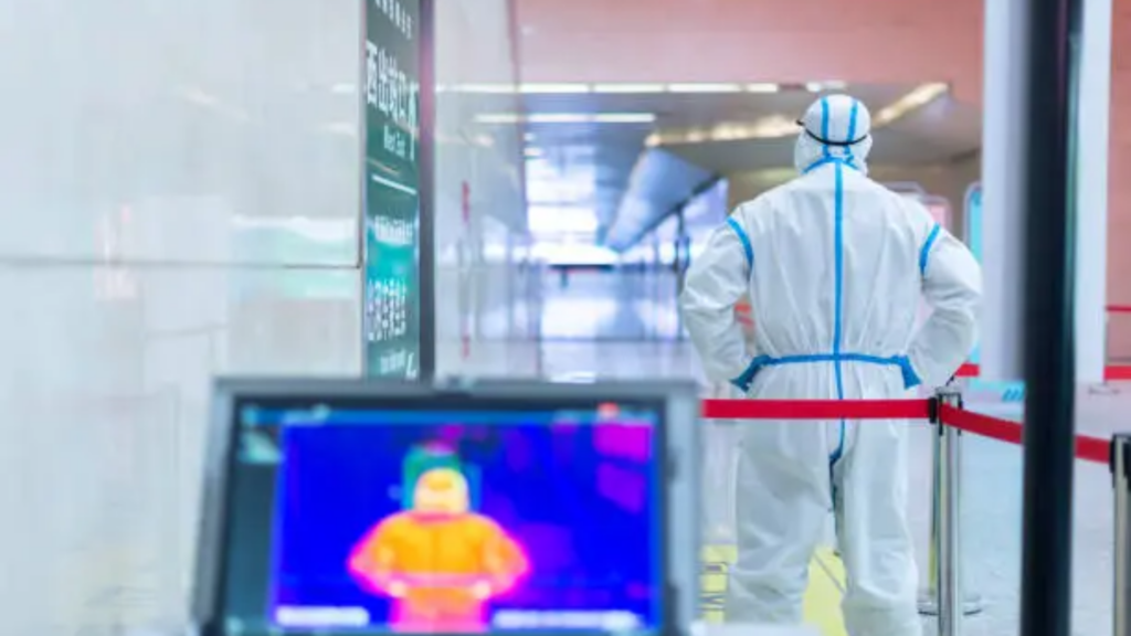 Person in protective gear standing near a thermal scanner in a medical facility.