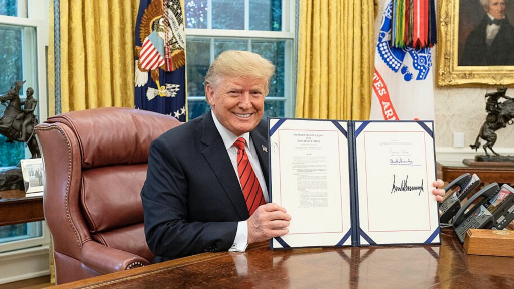 Donald Trump smiles while holding up a large signed document at his desk in the Oval Office.