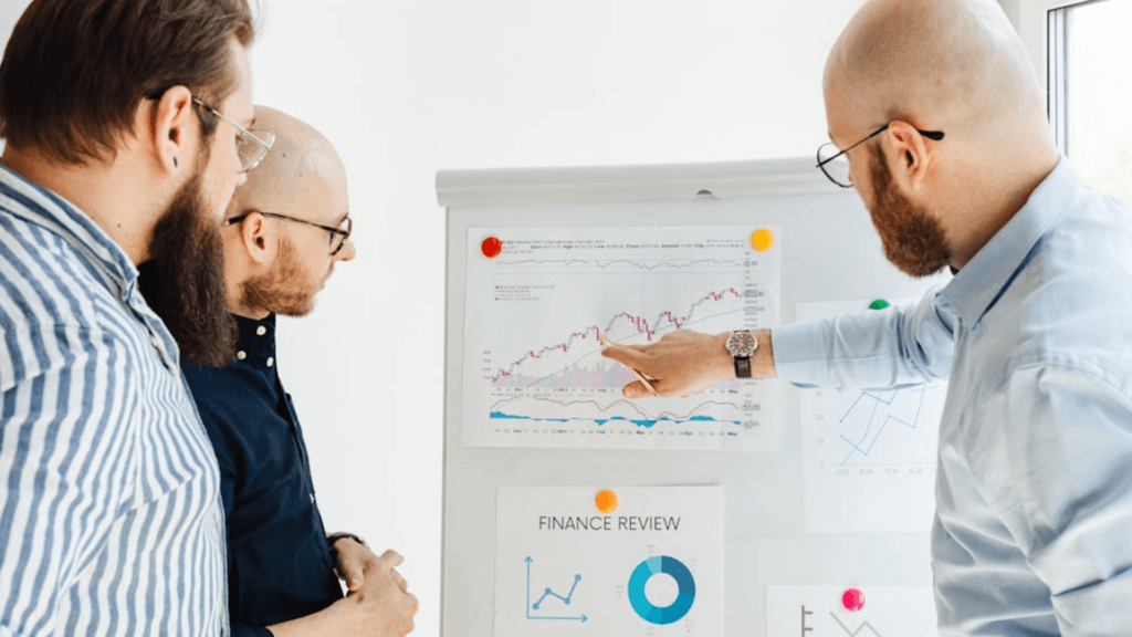 Three men stand together looking at an upward-trending financial line graph on a whiteboard.