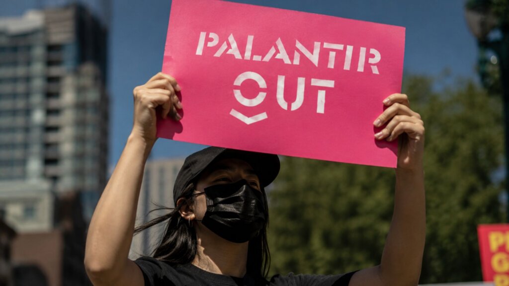 Person wearing a black hat and face mask holds a bright pink protest sign above their head that reads “PALANTIR OUT” during a daytime demonstration in an urban area with blurred buildings and trees in the background.