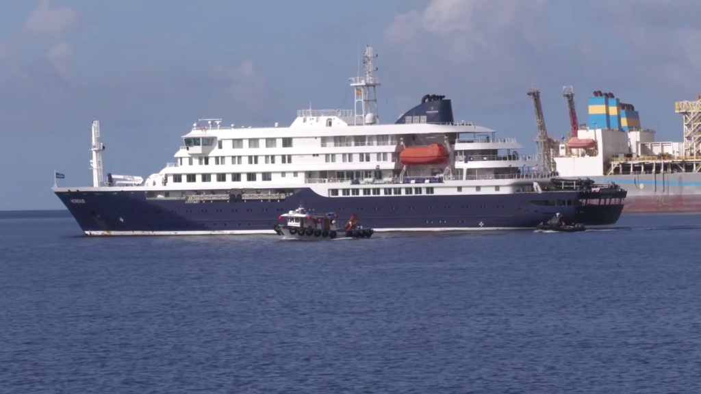 A large, dark blue and white passenger ship named "Hondius" travels across calm blue water under a hazy sky, accompanied by two smaller black support boats.