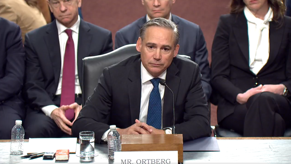 A man in a dark suit and blue tie, identified by a desk nameplate as Mr. Ortberg, speaks into a microphone while seated at a witness table during a government committee hearing.
