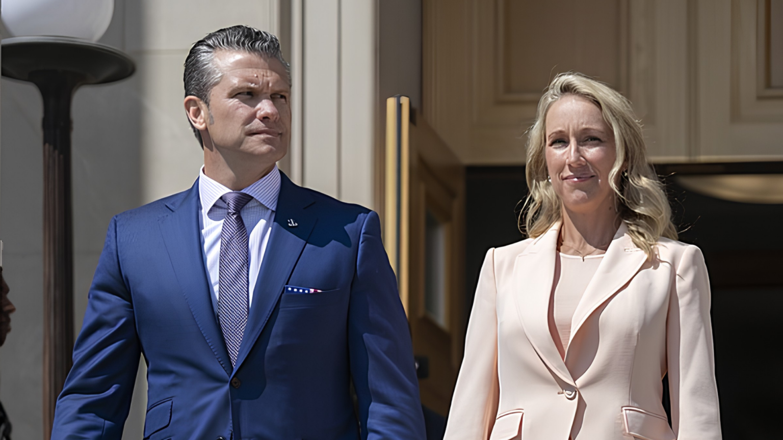 Secretary of War Pete Hegseth and his wife, Jennifer Rauchet, stand outside the entrance of a government building.