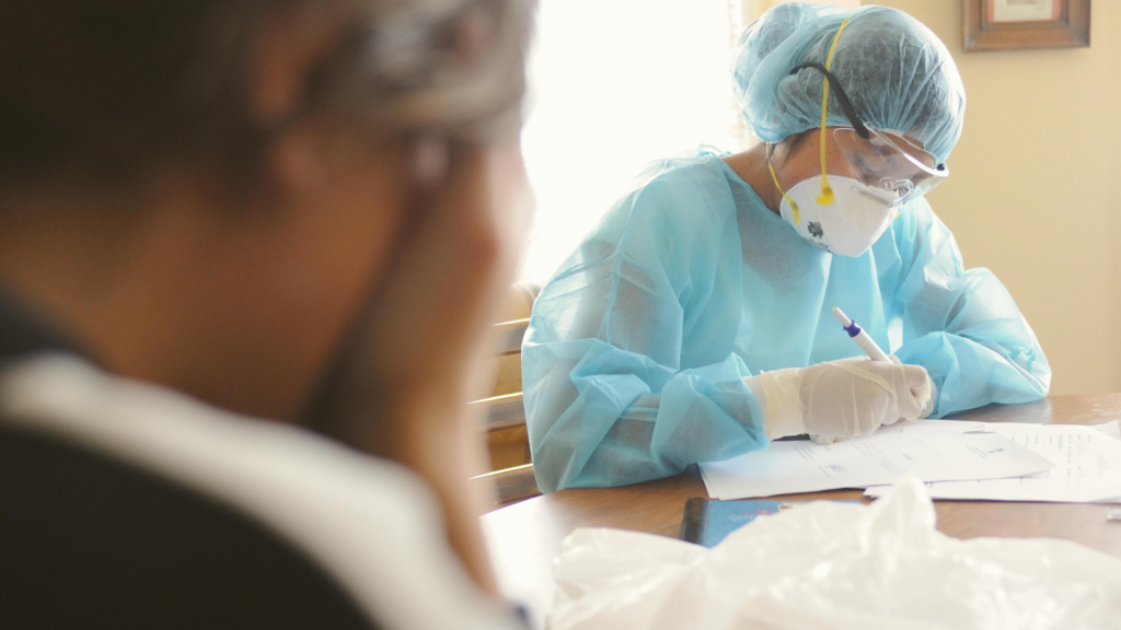 A close-up, angled view focusing on a person in a blue medical gown, safety goggles, and a respirator mask as they write on paperwork at a table.