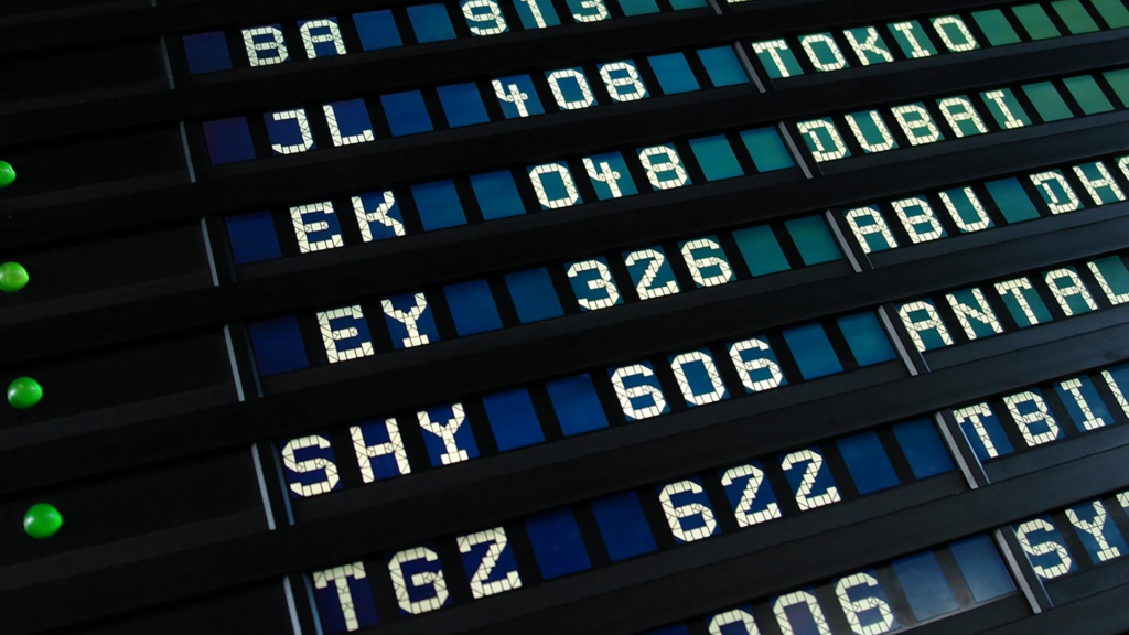 A close-up, low-angle shot of a mechanical split-flap airport departures board displaying various international flight codes and destinations.