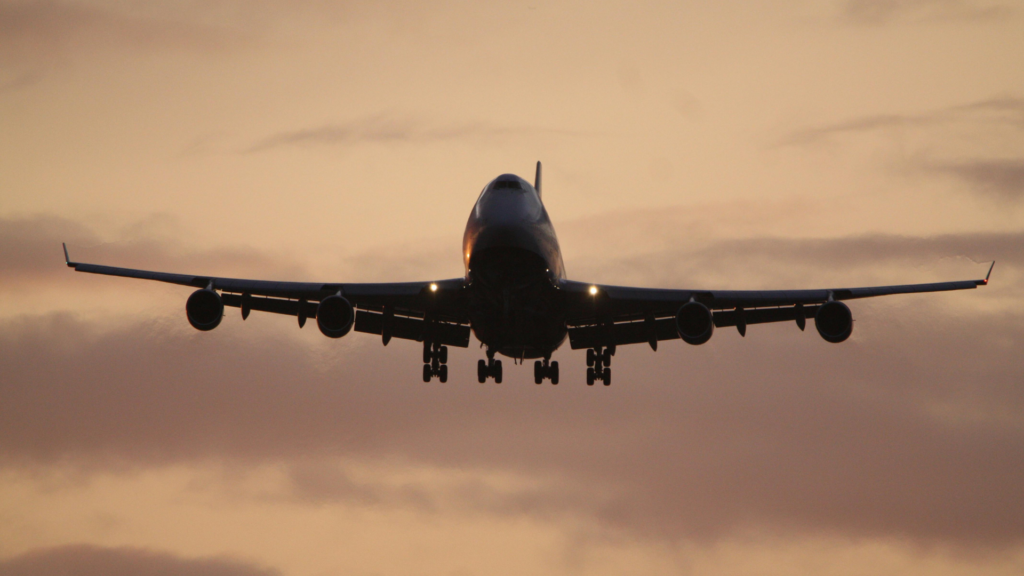 A front-facing silhouette of a large Boeing 747 airplane descending for landing with its landing gear down against a hazy, orange-tinted sunset sky.