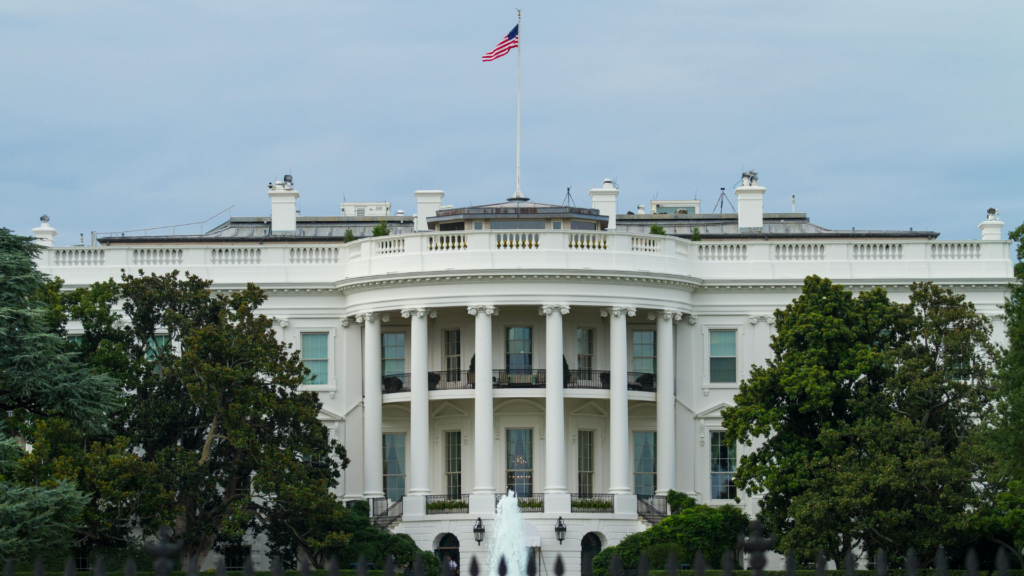 A straight-on view captures the South Portico of the White House under an overcast sky, framed by green trees and a black iron fence in the foreground.